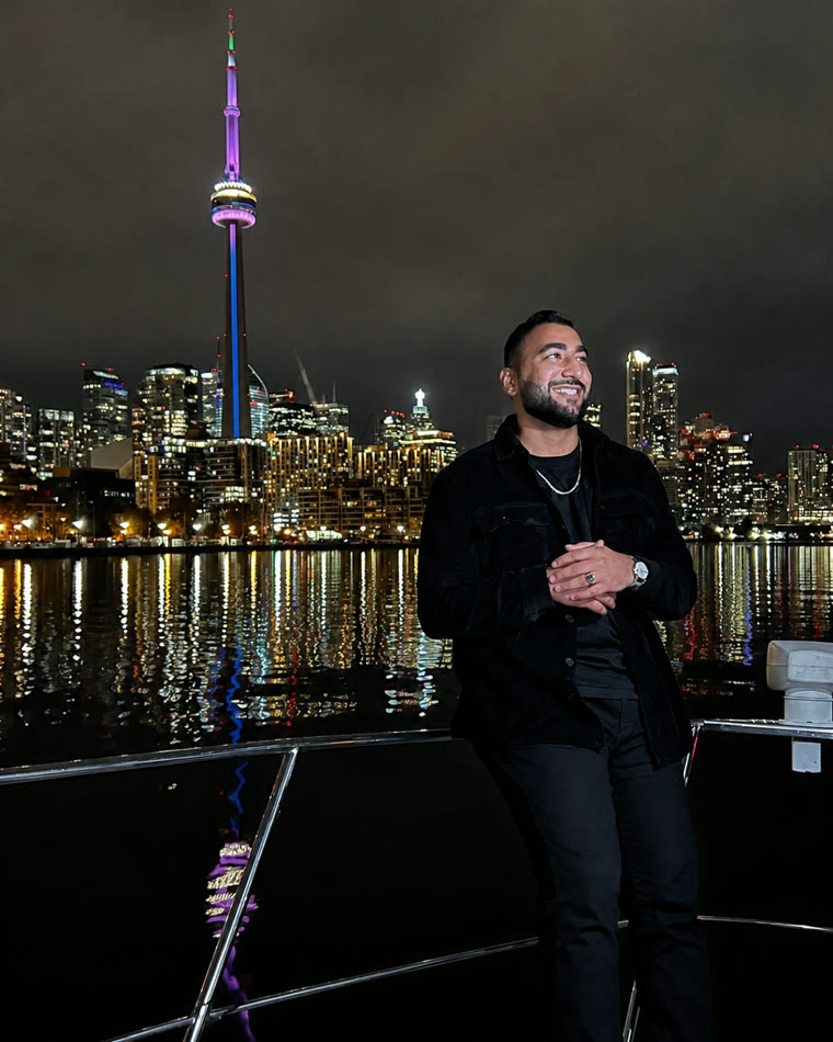 A man in black standing at the rail of a boat, the Toronto skyline and CN Tower lit behind him at night